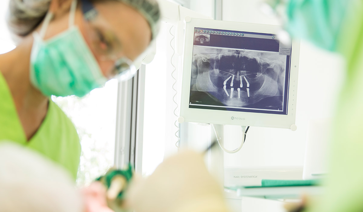 Mitarbeiterin mit Mundschutz beugt sich vornüber und hält Zahnarztinstrumente in der Hand, im Vordergrund ist eine andere Person mit Mundschutz zu sehen. Im Hintergrund sieht man ein Röntgenbild mit Zahnimplantaten.